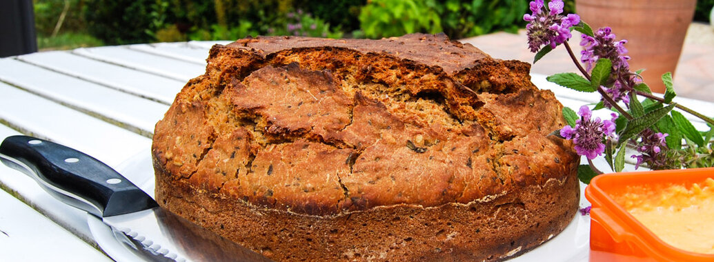 Frisch gebackenes Brot mit Knusprig-aufgebrochener Kruste, aus runder Backform. Dabeneben ein Schälchen mit gelben Kichererbsenaufstrich, ein Brotmesser und Violette Blumen
