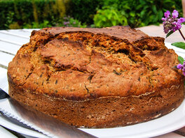 Frisch gebackenes Brot mit Knusprig-aufgebrochener Kruste, aus runder Backform. Dabeneben ein Schälchen mit gelben Kichererbsenaufstrich, ein Brotmesser und Violette Blumen