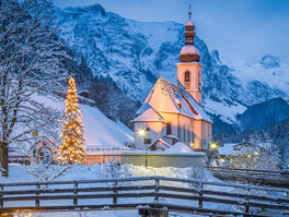 kleine angeleuchtete Kirche mit Weihnachtsbaum vor verschneiter Berglandschaft