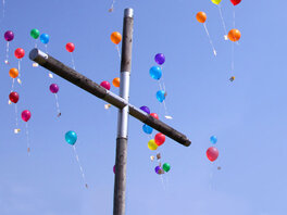 Hinter einem Kreuz, bunte zum Himmel austeigende Luftballons.