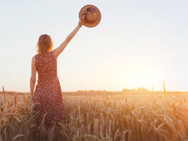 Frau mit Kleid im Kornfeld. Winkt mit einem Strohhut in Richtung Sonnenuntergang.