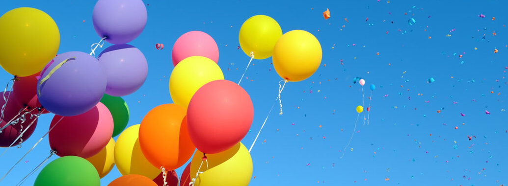Ein Bündel schwebende bunte Luftballons vor blauem Himmel.