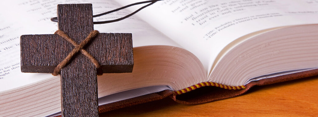 Schlichtes Holzkreuz an Stoffkette und ein geöffnetes Buch.