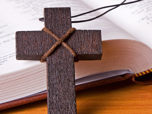 Schlichtes Holzkreuz an Stoffkette und ein geöffnetes Buch.