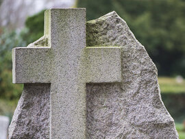 Grabstein mit in Stein gearbeitetes Kreuz. Im Hintergrund unscharf ein Friedhof.