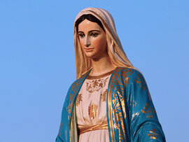 Marienfeste - Oberkörper einer Madonna mit Schleier, in einem rosa und blauen Gewand mit auffälligen Goldverzierungen vor klarem blauen Himmel.