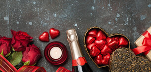 Zwei rote Rosen, ein Teelicht, eine Sektflasche, herzförmige Pralinen und Geschenk vor dunklem Hintergrund