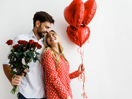 Junges Paar aneineinander gelehnt und sich anschauend. Er im weißen Hemdhält einen Strauß rote Rosen. Sie in roter Bluse hält drei rote Herzförmige Ballons.
