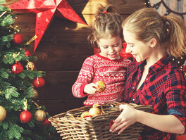 lächelnde Mutter mit Kind und Weidenkorb voller Christbaumkugeln auf dem Schoß neben geschmücktem Weihnachtsbaum