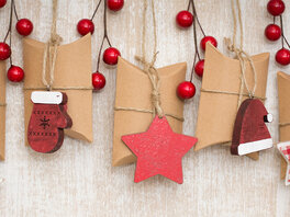 Ein Adventskalender mit schlichten Pappschachteln einzeln an Schnüren mit dekorativen Anhängern sowie roten Stechpalmenzweigen.