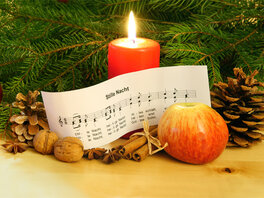 Notenblatt "Stille Nacht". Dekoriert mit Tannengrün, brennender Kerze, Apfel, Nüssen, Zapfen, Kugeln.