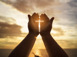 Geöfnette Hände einer Person, der Sonne im Dämmerlicht entgegen gestreckt. Zwischen den Händen scheint die Sonne hindurch und bildet ein Lichtkreuz.