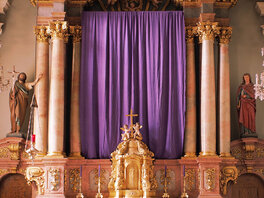 Hochaltar, dessen Mittelteil mit einem violettem Fastentuch abgedeckt ist. Der Altar ist mit kunstvollen Säulen, Figuren und Ornamenten gestaltet.