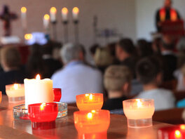 Gottesdienst in der Kirche. Im Vordergrund stehen brennende Kerzen, im Hintergrund ist der Altar mit Kreuz mit brennden Kerzen, der Pfarrer und die Gemeinde zu sehen.