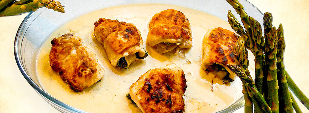 Knusprig-goldene Putenröllchen in heller Soße in einer ovalen Glasauflaufform. Dazu grüner Spargel.