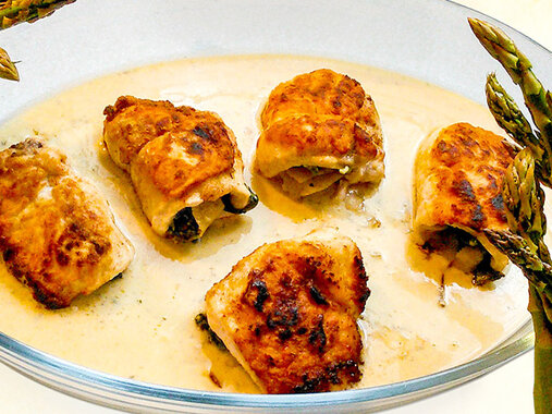 Knusprig-goldene Putenröllchen in heller Soße in einer ovalen Glasauflaufform. Dazu grüner Spargel.
