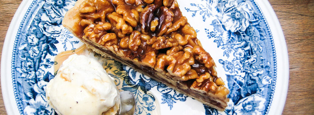 Stück Walnusstarte bedecktmit glänzender Wallnuss-Karamell-Schicht, dazu ein Kugel cremiges Vanilleeis mit zerfließend-goldigem Ahornsirup.