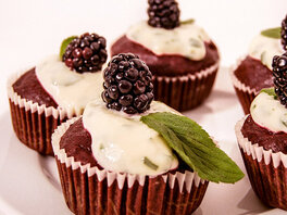 Fünf kräftig rot-braune Muffins in Papierförmchen, bedeckt mit feiner Pfefferminzcreme, einem frischen Minzblatt, sowie je einer kräftigen Brombeere obenauf.