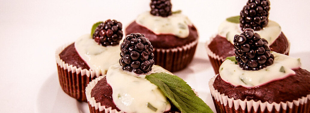 Fünf kräftig rot-braune Muffins in Papierförmchen, bedeckt mit feiner Pfefferminzcreme, einem frischen Minzblatt, sowie je einer kräftigen Brombeere obenauf.