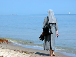 Ordenseintritt - Nonne geht mit Schuhen in den Händen am Ufer eines Meeres entlang