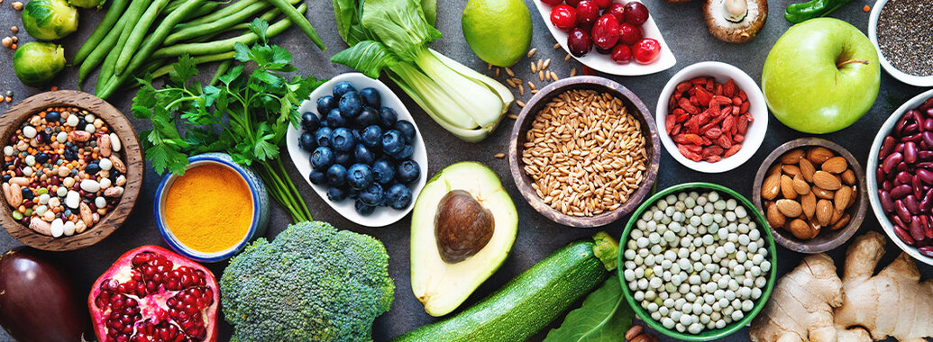 Auswahl an Lebensmitteln: Linsen, Bohnen, Aubergine, Rosenkohl, Granatapfel, grüne Bohnen, Brokkoli, Petersilie, Heidelbeeren, Pak Choi, Avocado, Zitrone, Getreide, Zucchini, Cranberries, Mandeln, Ingwer, Gojibeeren, Champignons, Chiasamen, Apfel, Kürbiskerne