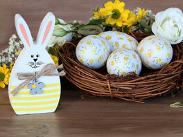 Nest mit Ostereiern, mit feinen gelben Blumen bemalt. Eiförmiger Osterhase, aus Holz. Gelbe Blumen.